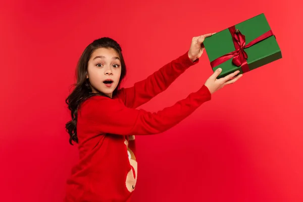 Schockiertes Mädchen im Winterpullover hält verpacktes Weihnachtsgeschenk auf rotem Grund — Stockfoto