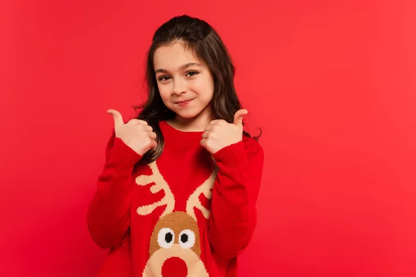 Positive girl in winter sweater showing thumbs up and looking at camera isolated on red — Stock Photo