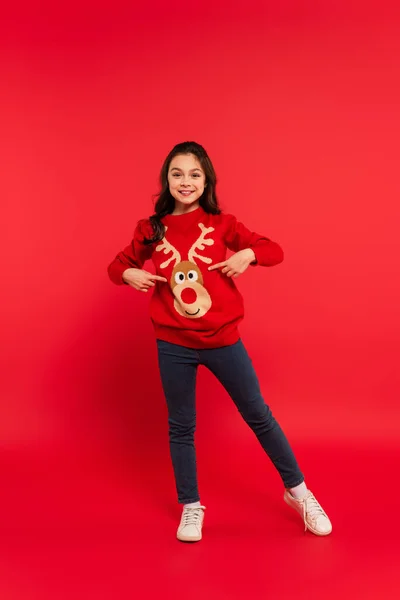Full length of cheerful girl pointing with fingers at Christmas sweater on red — Stock Photo