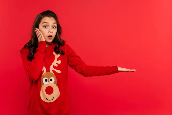 Surprised child in Christmas sweater pointing with hand isolated on red — Stock Photo
