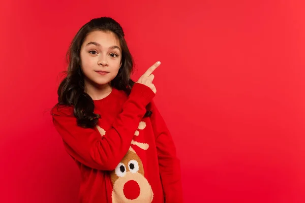 Cheerful kid in Christmas sweater pointing with finger isolated on red — Stock Photo