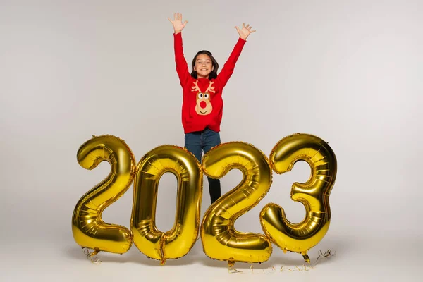 Pleine longueur d'enfant gai en pull rouge debout avec les mains levées près des ballons avec des numéros 2023 sur gris — Photo de stock