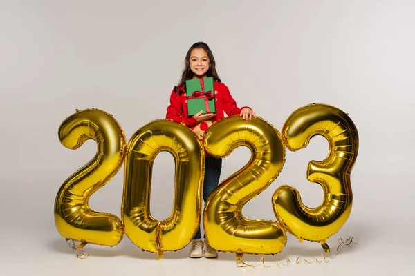 Full length of happy kid in  sweater holding christmas present near balloons with 2023 numbers on grey — Stock Photo