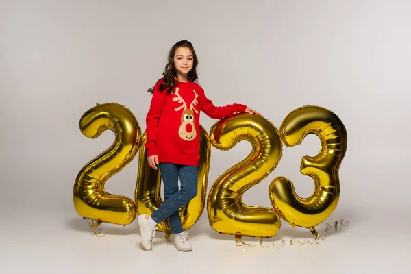 Full length of cheerful child in red sweater standing near balloons with 2023 numbers on grey — Stock Photo