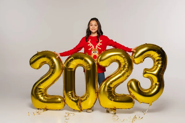 Pleine longueur de fille souriante en pull rouge debout près des ballons avec des numéros 2023 sur gris — Photo de stock