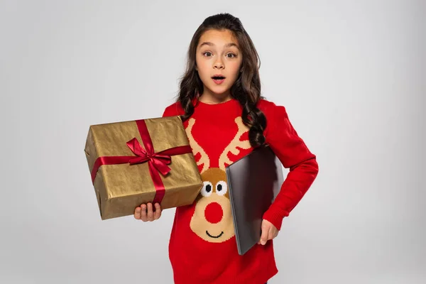 Shocked child in red sweater holding laptop and Christmas present on grey — Stock Photo
