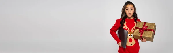 Enfant choqué en pull rouge tenant ordinateur portable et cadeau de Noël sur gris, bannière — Photo de stock