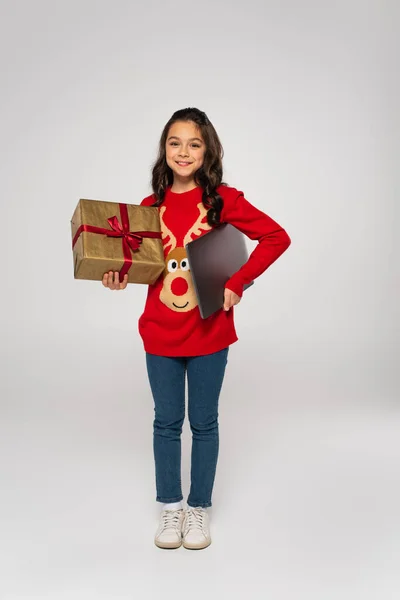 Longitud completa de la muchacha feliz en suéter rojo que sostiene el ordenador portátil y regalo de Navidad en gris - foto de stock