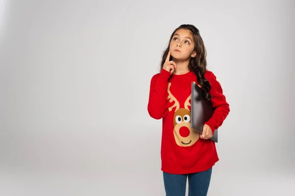 Niño pensativo en suéter rojo de Navidad que sostiene el ordenador portátil aislado en gris - foto de stock