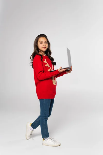 Full length of smiling child in red Christmas sweater holding laptop on grey — Stock Photo