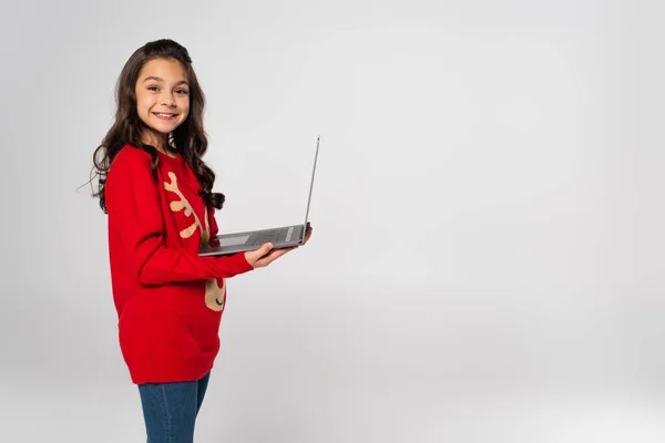 Menina feliz em camisola de Natal vermelho segurando laptop isolado no cinza — Fotografia de Stock