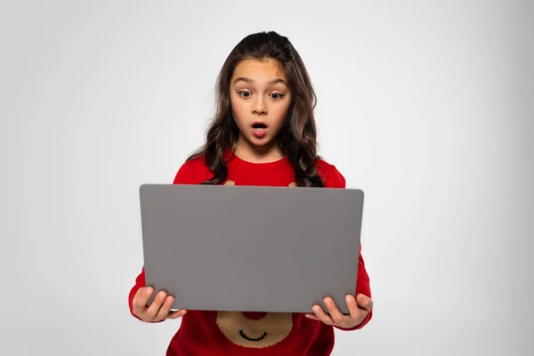 Шокована дитина в червоному різдвяному светрі дивиться на ноутбук ізольовано на сірому — стокове фото