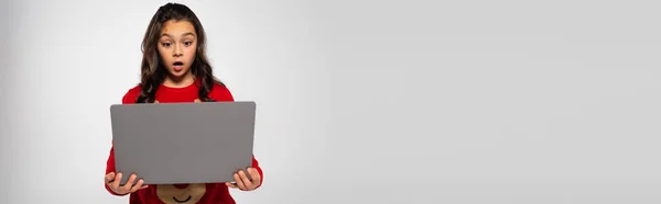 Shocked child in red knitted sweater looking at laptop isolated on grey, banner — Stock Photo