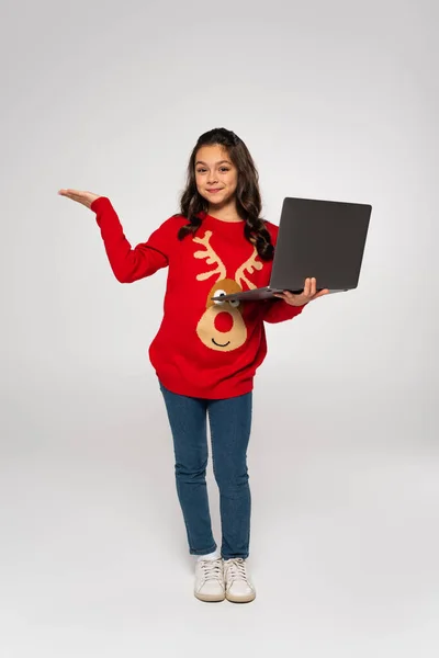 Pleine longueur de joyeux enfant en pull rouge tenant ordinateur portable et pointant avec la main sur le gris — Photo de stock