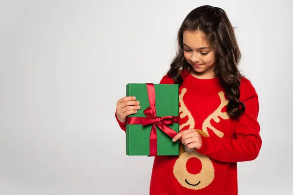Glückliches Mädchen im roten Pullover, das verpackte Weihnachtsgeschenk isoliert auf grau betrachtet — Stockfoto