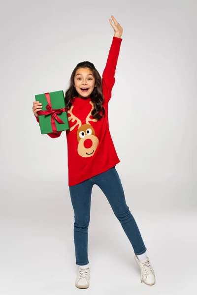 Full length of amazed girl in red sweater holding Christmas present on grey — Stock Photo