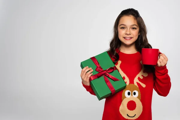 Glückliches Mädchen in rotem Pullover mit Tasse Kakao und Weihnachtsgeschenk isoliert auf grau — Stockfoto