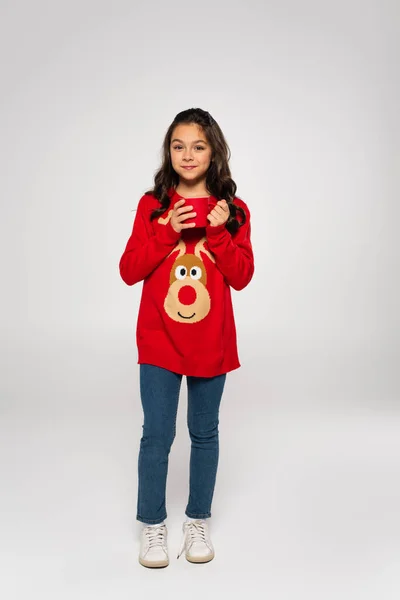 Полная длина счастливой девушки в красном свитере, держащей чашку коки на сером — стоковое фото