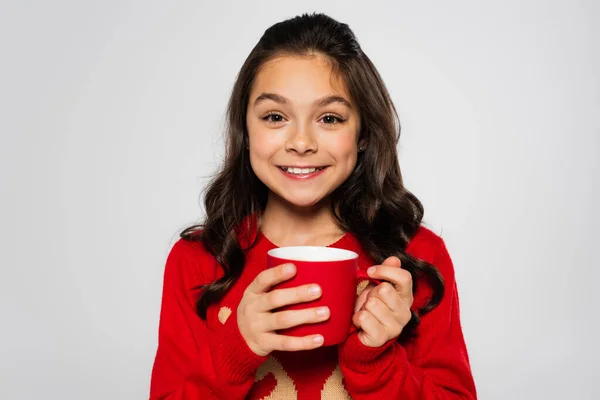 Cheerful girl in red sweater holding cup and looking at camera isolated on grey — Stock Photo