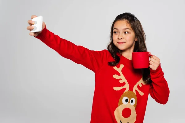 Smiling girl in New Year sweater holding cup and taking selfie isolated on grey — Stock Photo