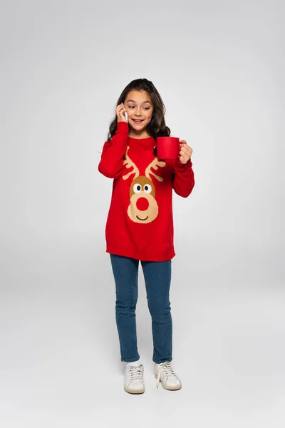 Полная длина ребенка в новогоднем свитере, говорящего по смартфону и держащего чашку на сером — стоковое фото