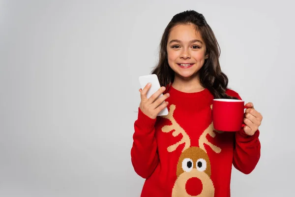 Позитивный ребенок в новогоднем свитере с кубком и смартфоном, изолированным на сером — стоковое фото
