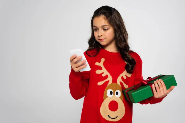 Preteen girl in Christmas sweater holding gift and using smartphone isolated on grey — Stock Photo