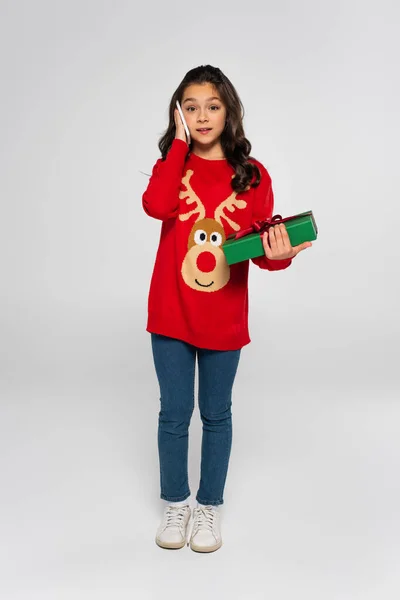 Longitud completa de la chica en suéter hablando en el teléfono inteligente y la celebración de regalo de Navidad en gris - foto de stock