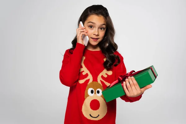 Preteen girl in sweater talking on smartphone and holding Christmas gift isolated on grey — Stockfoto