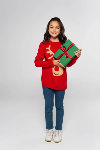 Lunghezza completa della ragazza allegra in maglione rosso lavorato a maglia con regalo su sfondo grigio — Foto stock