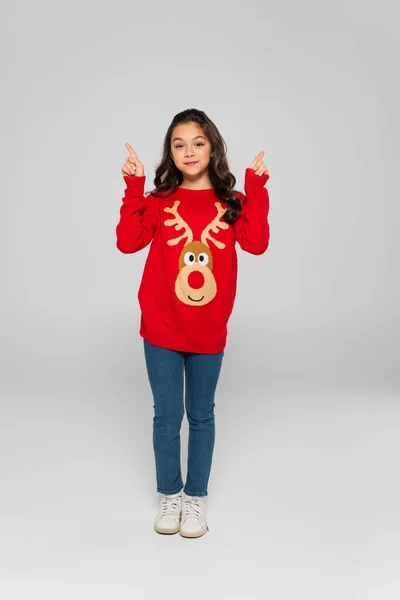 Повна довжина дитини в новорічному светрі, що вказує пальцями на сірий фон — стокове фото