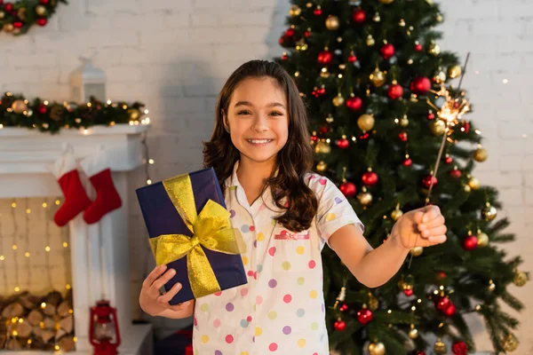 Fröhliches Mädchen im Pyjama hält Geschenk und Wunderkerze neben Weihnachtsbaum zu Hause — Stockfoto