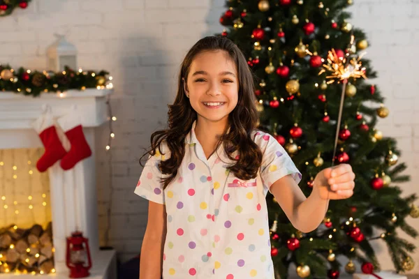 Улыбающаяся девушка в пижаме держит искру и смотрит в камеру во время празднования Рождества дома — стоковое фото