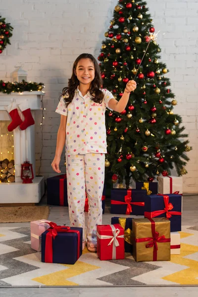 Lächelndes Kind im Pyjama hält Wunderkerze in der Nähe von Geschenken und Weihnachtsbaum zu Hause — Stockfoto