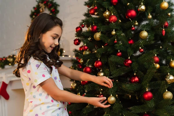 Vista lateral da menina no pijama segurando bola perto da árvore de Natal em casa — Fotografia de Stock