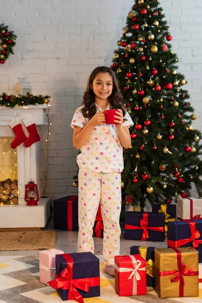 Bambino sorridente in pigiama che tiene la tazza vicino all'albero di Natale e i regali a casa — Foto stock