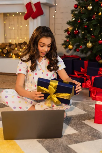 Muchacho emocionado en pijama punteada apertura caja de regalo cerca de la computadora portátil y el árbol de Navidad en casa - foto de stock