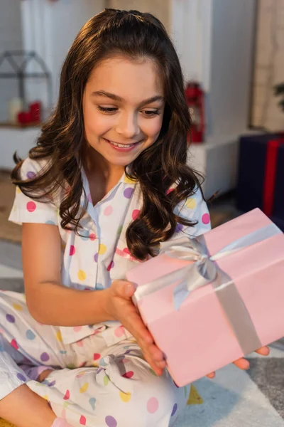 Enfant souriant en pyjama pointillé tenant une boîte-cadeau floue pendant la célébration de Noël à la maison — Photo de stock