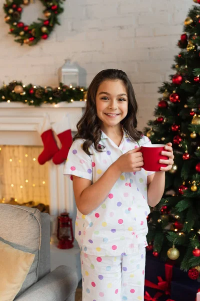 Niño positivo en pijama sosteniendo la taza cerca del árbol de Navidad en casa por la noche - foto de stock
