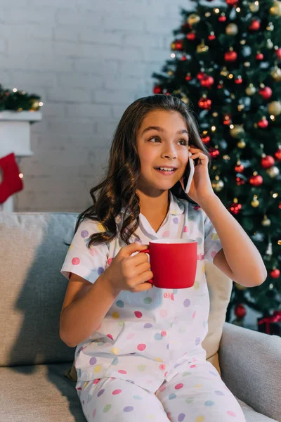 Ребенок-подросток в пижаме разговаривает на смартфоне и держит кубок во время празднования Рождества дома — стоковое фото