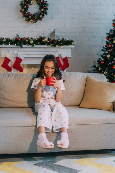 Lächelndes Mädchen im Pyjama, das eine Tasse hält, während es zu Hause auf der Couch neben dem Weihnachtsbaum sitzt — Stockfoto