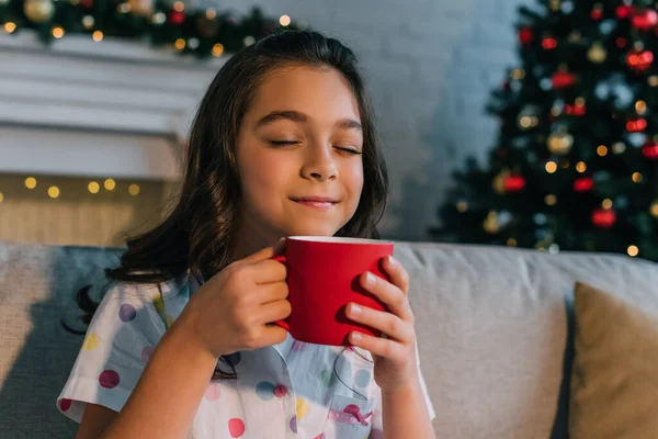 Gefallen preteen Kind im Pyjama riechendes Getränk in Tasse während Weihnachtsfeier zu Hause — Stockfoto