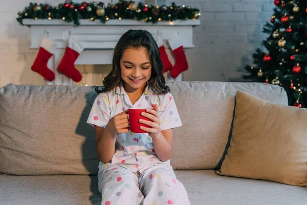 Cheerful girl in dotted pajama holding cup on couch near Christmas tree at home — Stock Photo