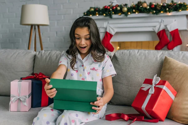 Excited kid in pajama opening present on couch during Christmas celebration at home — Stock Photo