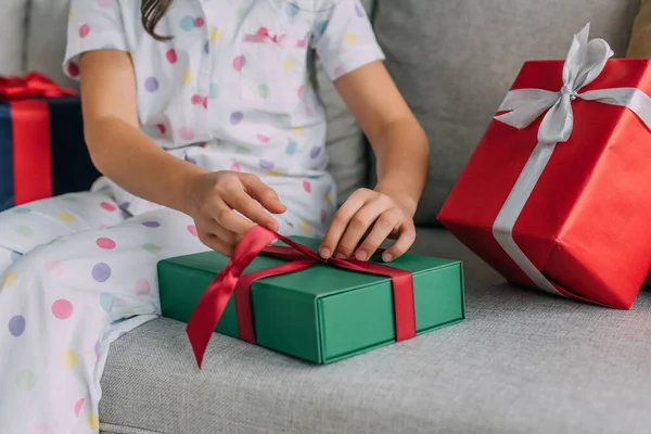 Cropped view of preteen kid in pajama untying ribbon of Christmas gift on couch — Stock Photo
