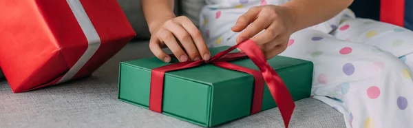 Cropped view of child in pajama untying bow on Christmas gift on couch, banner — Stock Photo