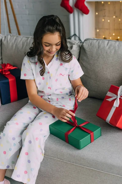 Счастливый ребенок в пижаме развязывает ленточку на подарок во время празднования Рождества дома — стоковое фото