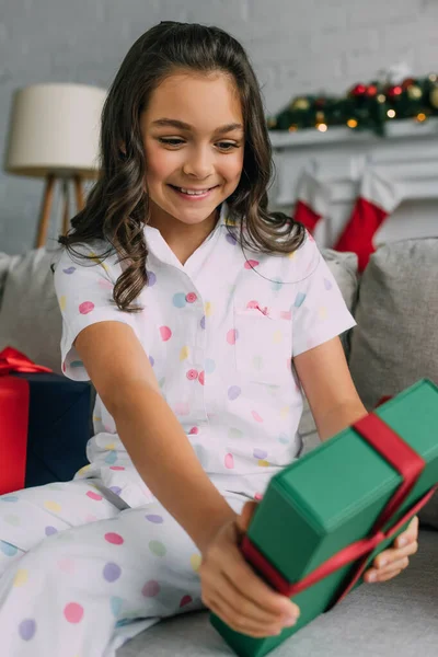 Happy girl in dotted pajama holding blurred Christmas gift on couch at home — Stock Photo