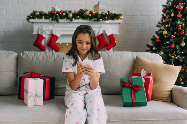Menina no pijama usando celular perto de presentes de Natal e pinheiros decorados em casa — Fotografia de Stock