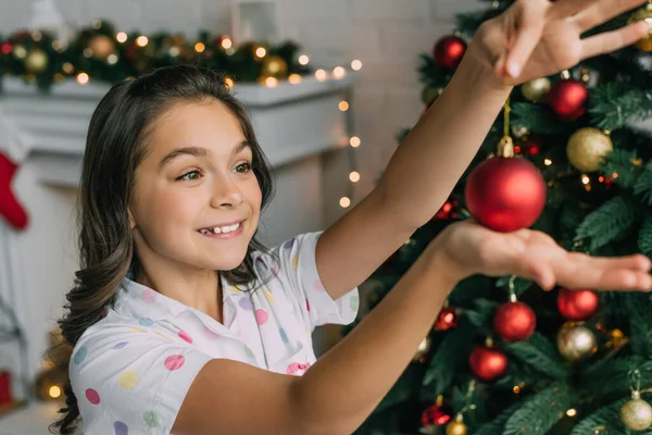 Chica alegre en pijama sosteniendo bola borrosa cerca del árbol de Navidad en casa - foto de stock